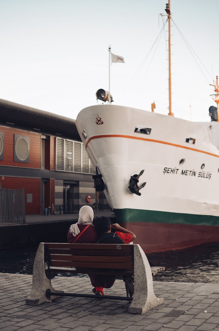 Two Person Sitting On Bench Facing Boat On Dock