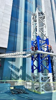 Workers on a crane assembling a skyscraper under construction against a glass facade.