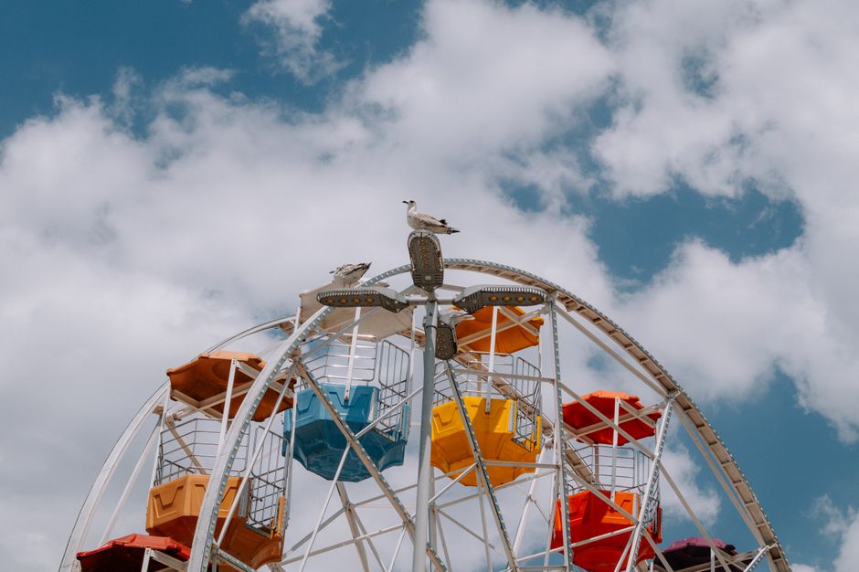 A vibrant ferris wheel with seagulls perched on top, set against a bright cloudy sky.