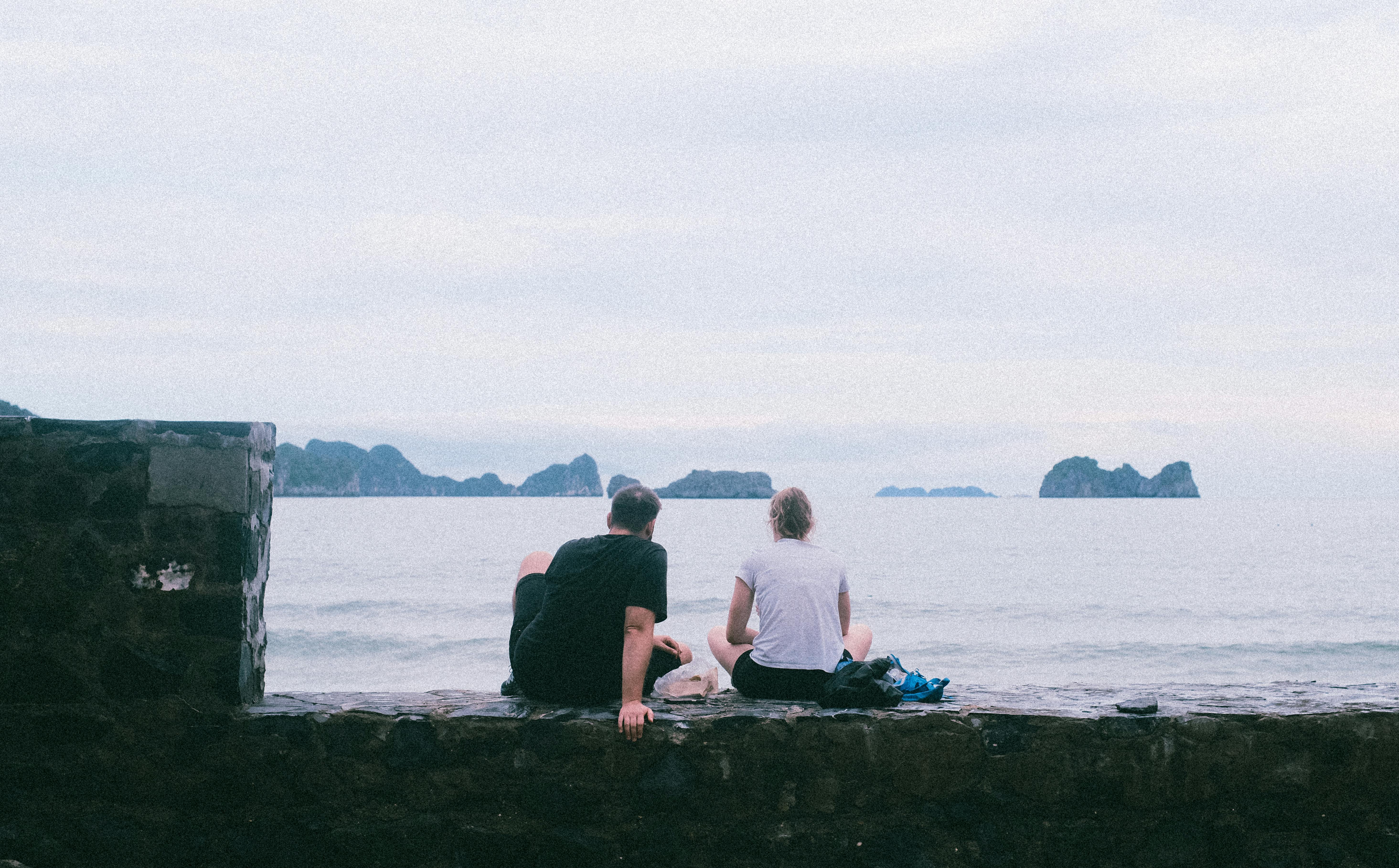 two people sitting on a wall looking out to the ocean