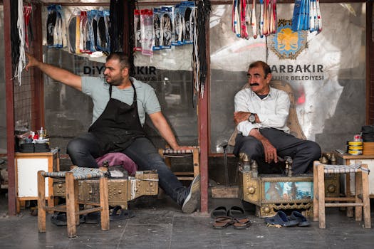 Two men sit and relax in a shoeshine stall at a Diyarbakır market, displaying products.