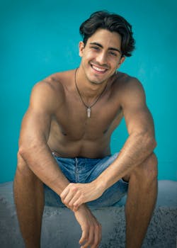 Shirtless young man smiling in shorts with blue background, embodying summer vibes and casual style.