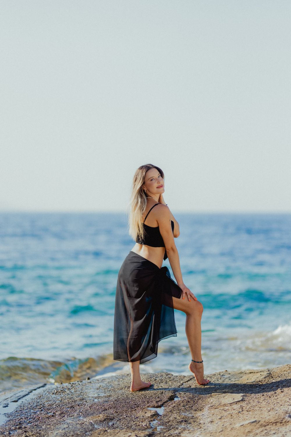 A woman in a black skirt and top is standing on a rock near the ocean ...