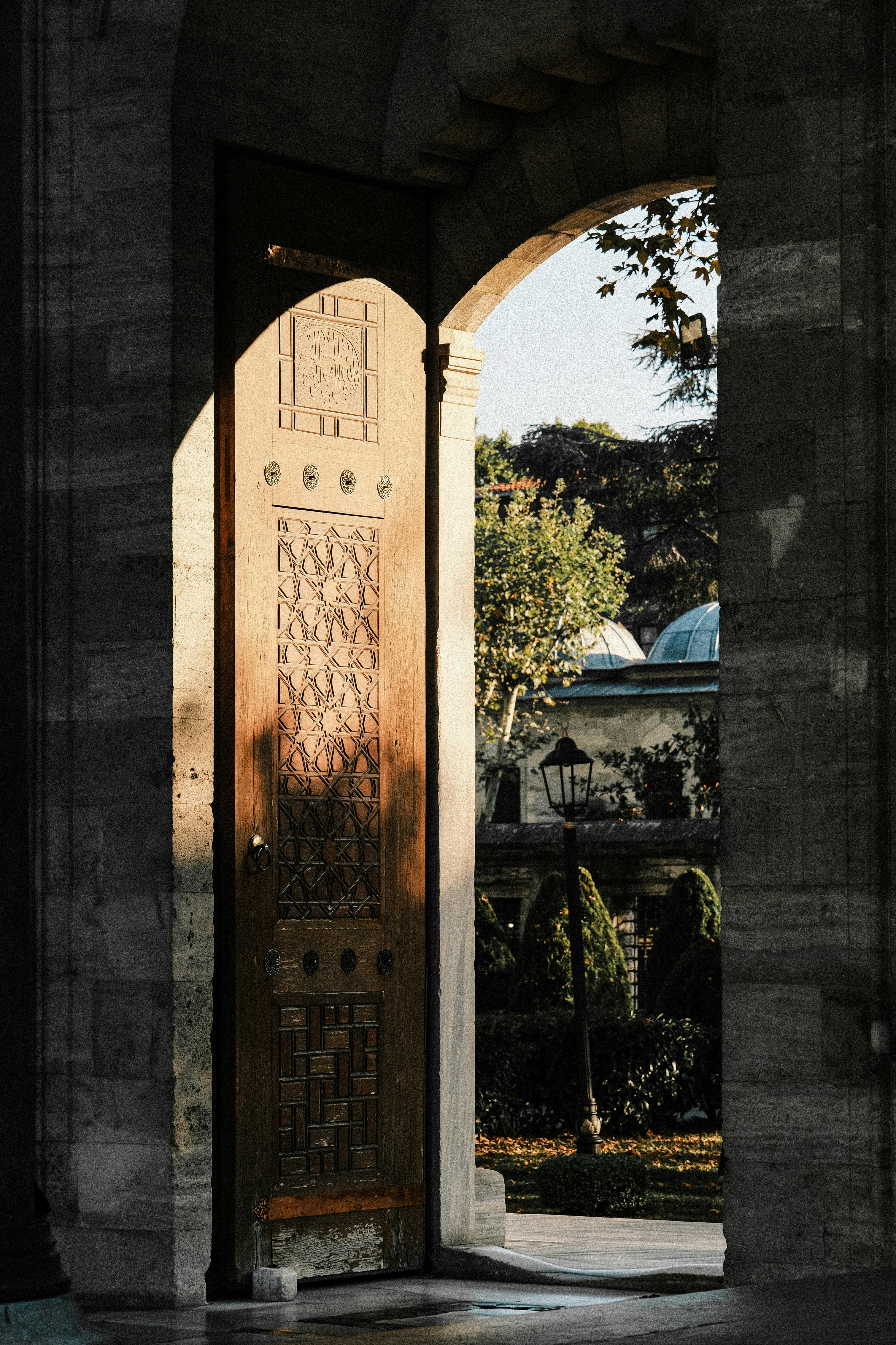 An intricately carved wooden door in a sunlit stone archway leads to a tranquil courtyard.