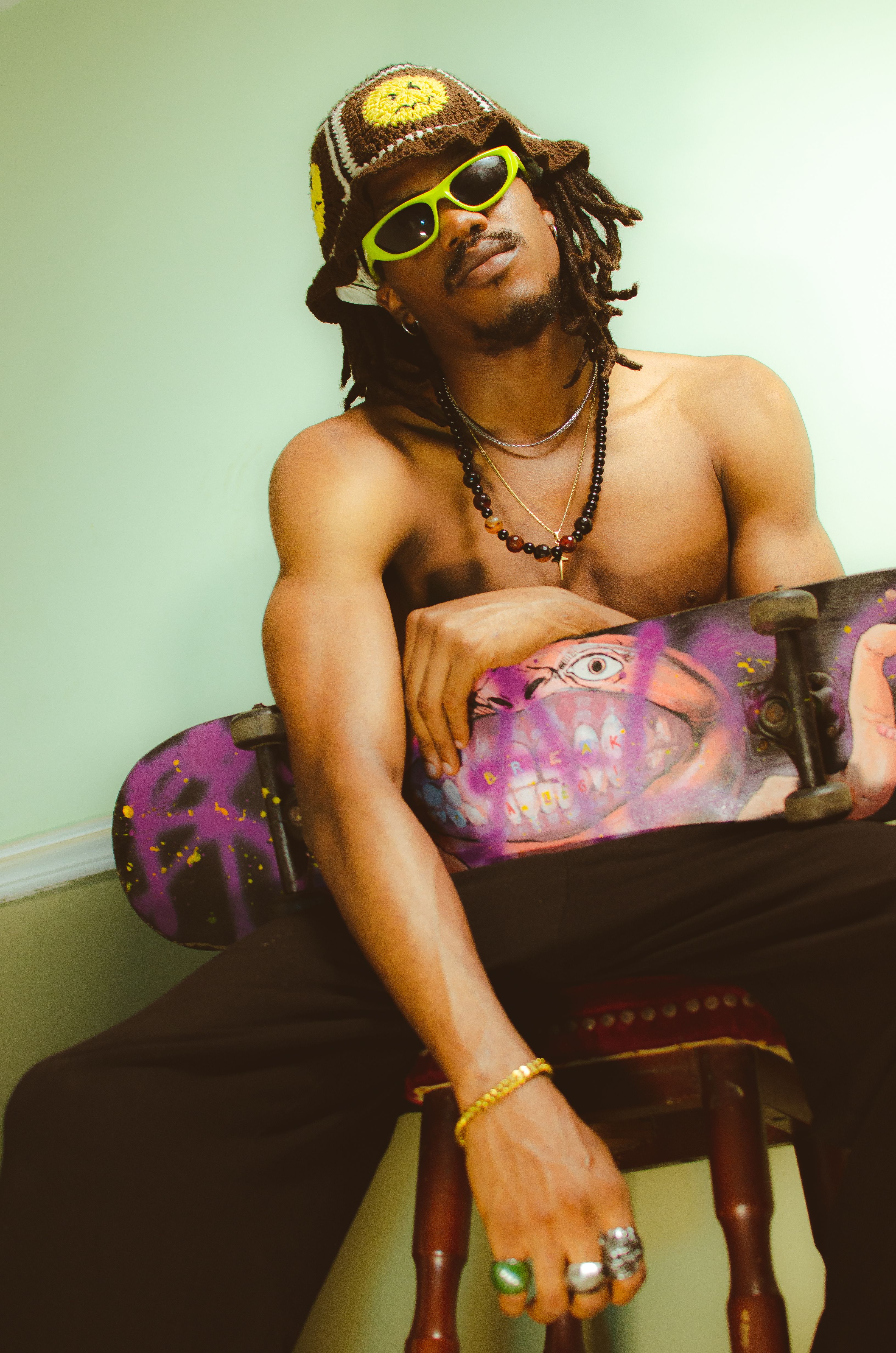 Shirtless skateboarder with sunglasses and accessories posing in a relaxed setting.
