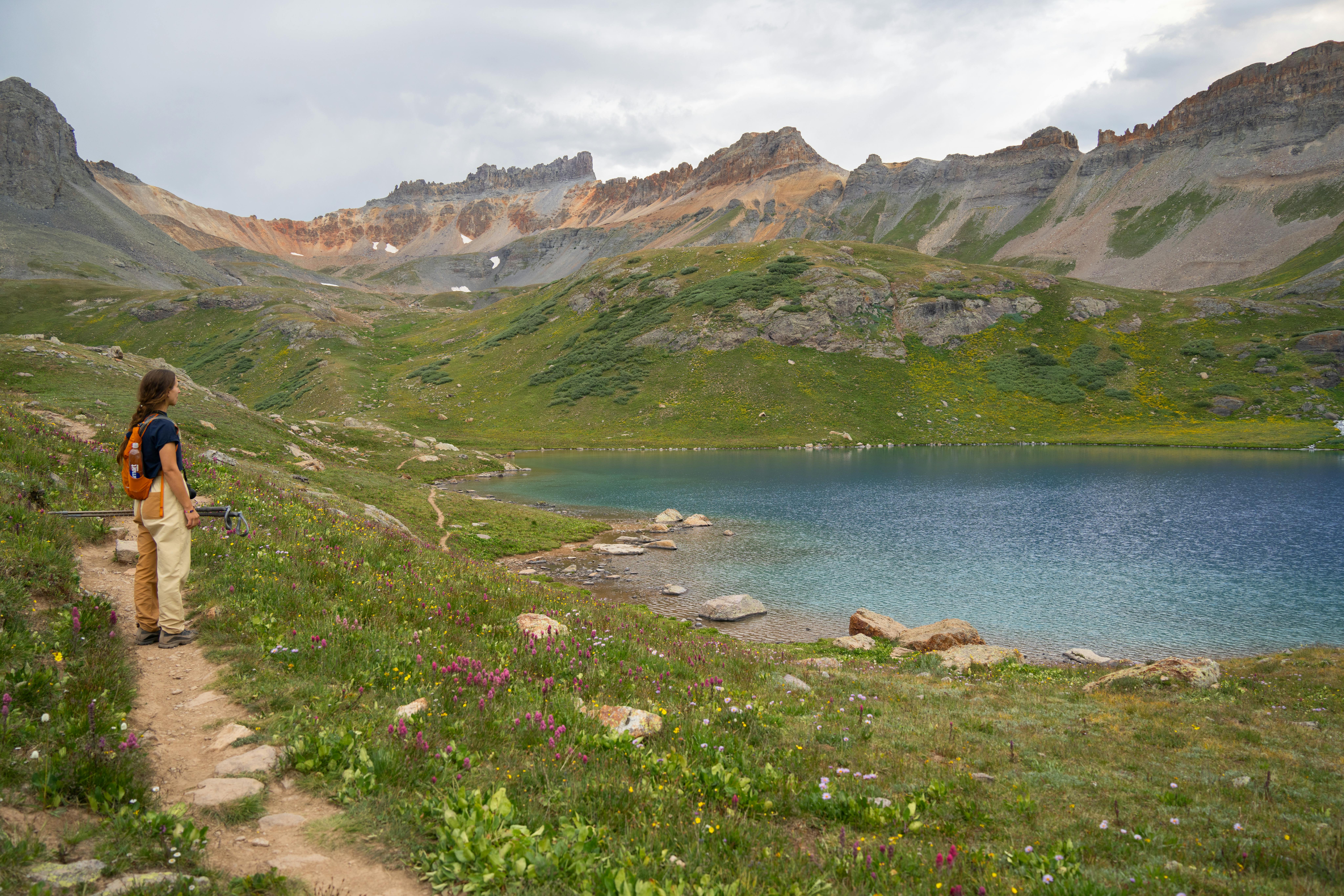 A woman hikes along a mountain trail by a serene blue lake surrounded by lush greenery.