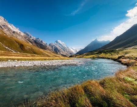Pristine turquoise river cutting through a valley with majestic snow-capped mountains under a clear blue sky.