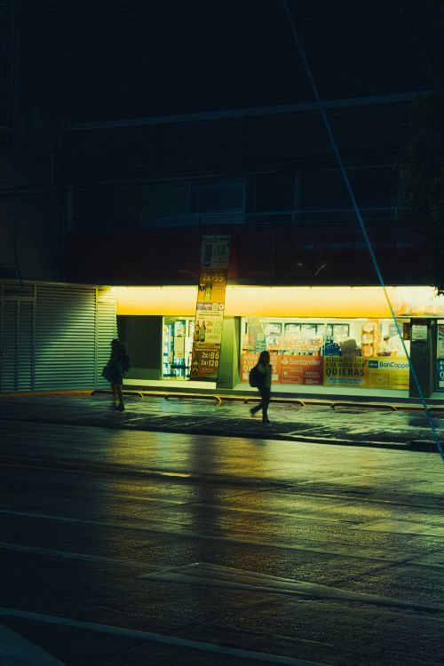 Free Moody urban night scene with pedestrians walking by an illuminated storefront, highlighting city life. Stock Photo