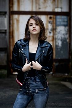 Fashion portrait of a young woman in a leather jacket and jeans posing confidently outdoors.