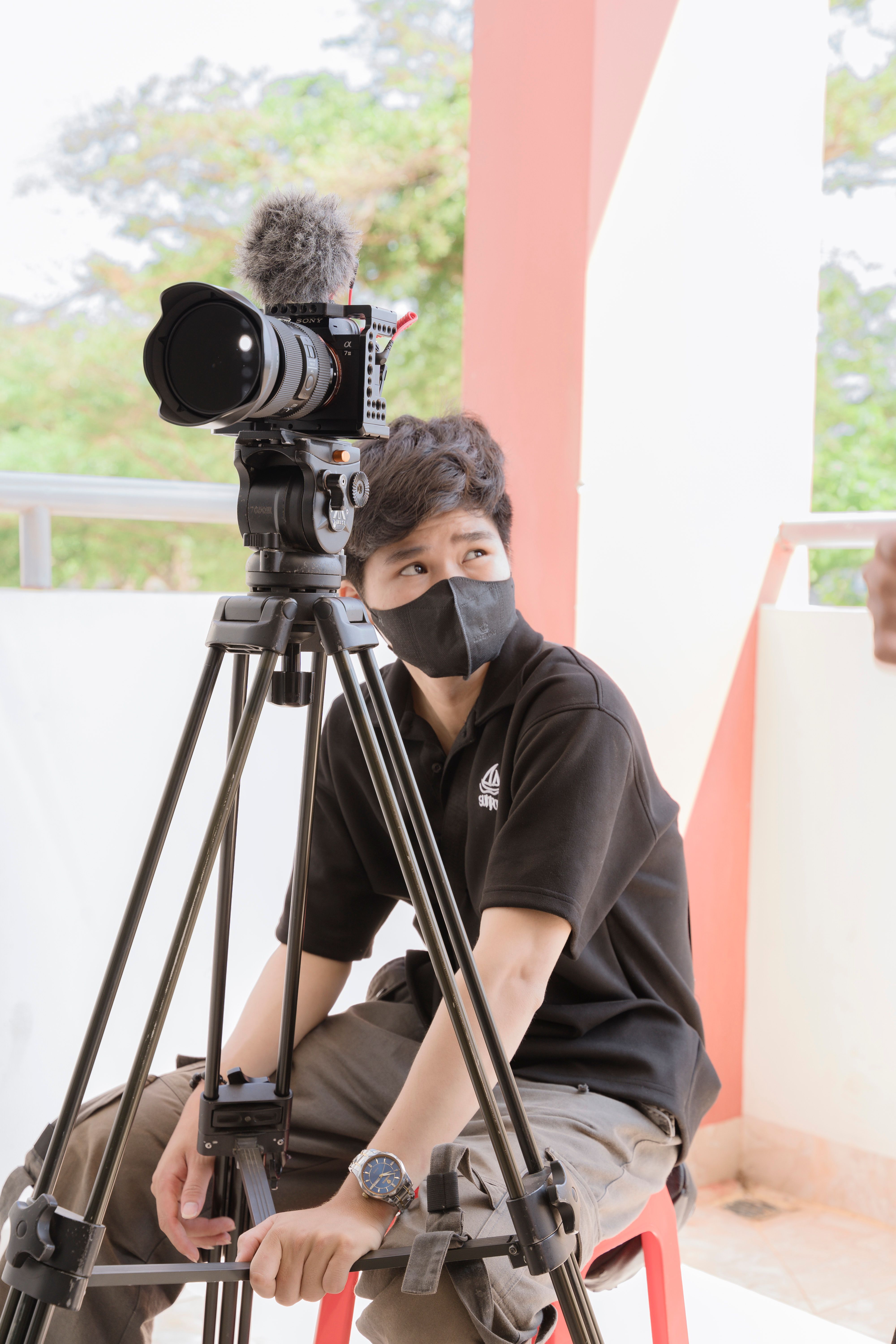 A young videographer sits outdoors, adjusting a camera on a tripod.