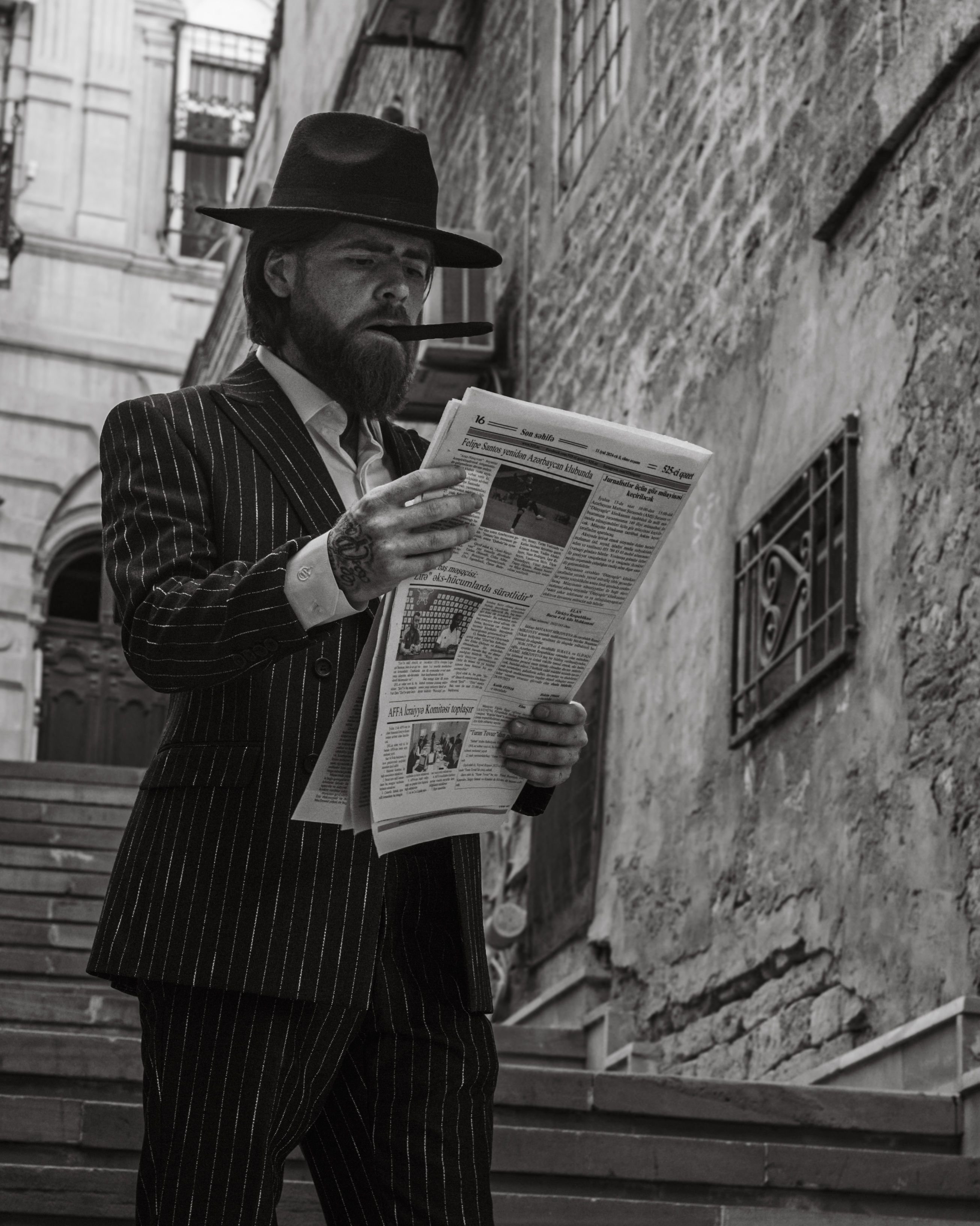 A stylish man in a pinstripe suit and fedora reads a newspaper on an old street in Baku, Azerbaijan.