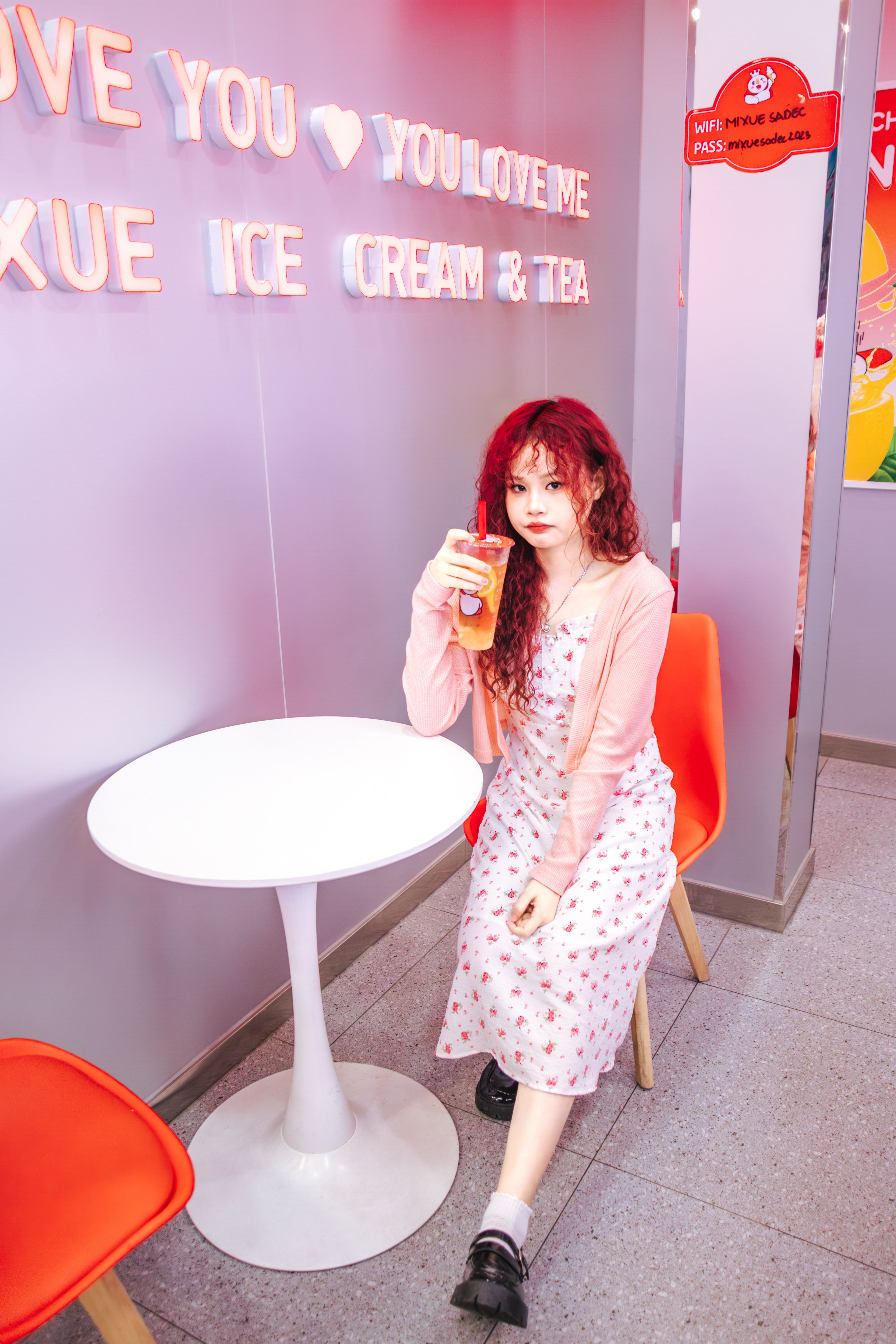 A stylish young woman with red hair enjoys a drink at a vibrant, modern cafe with neon signs.