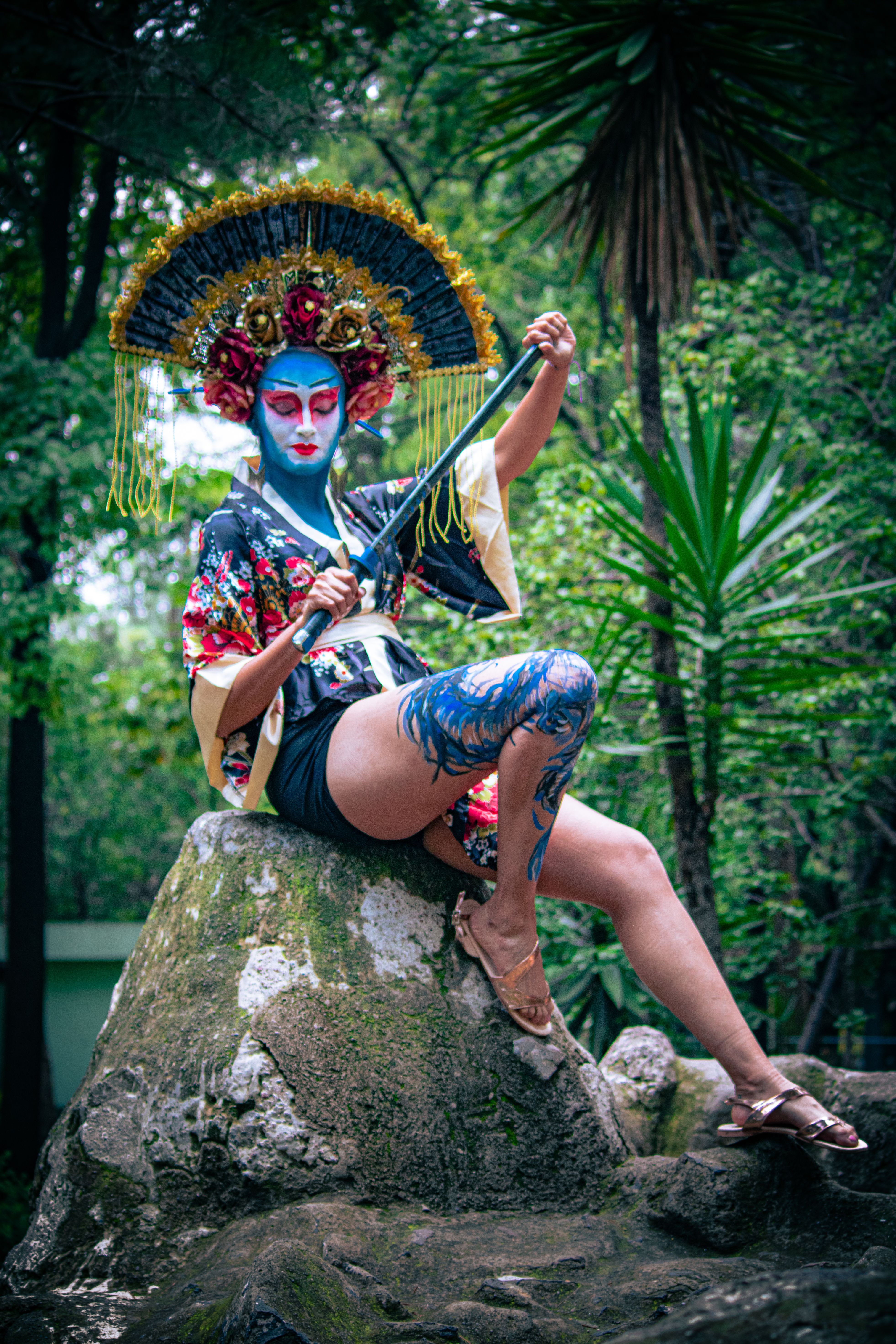 A person in dramatic geisha-inspired attire poses on a rock in a lush outdoor setting.