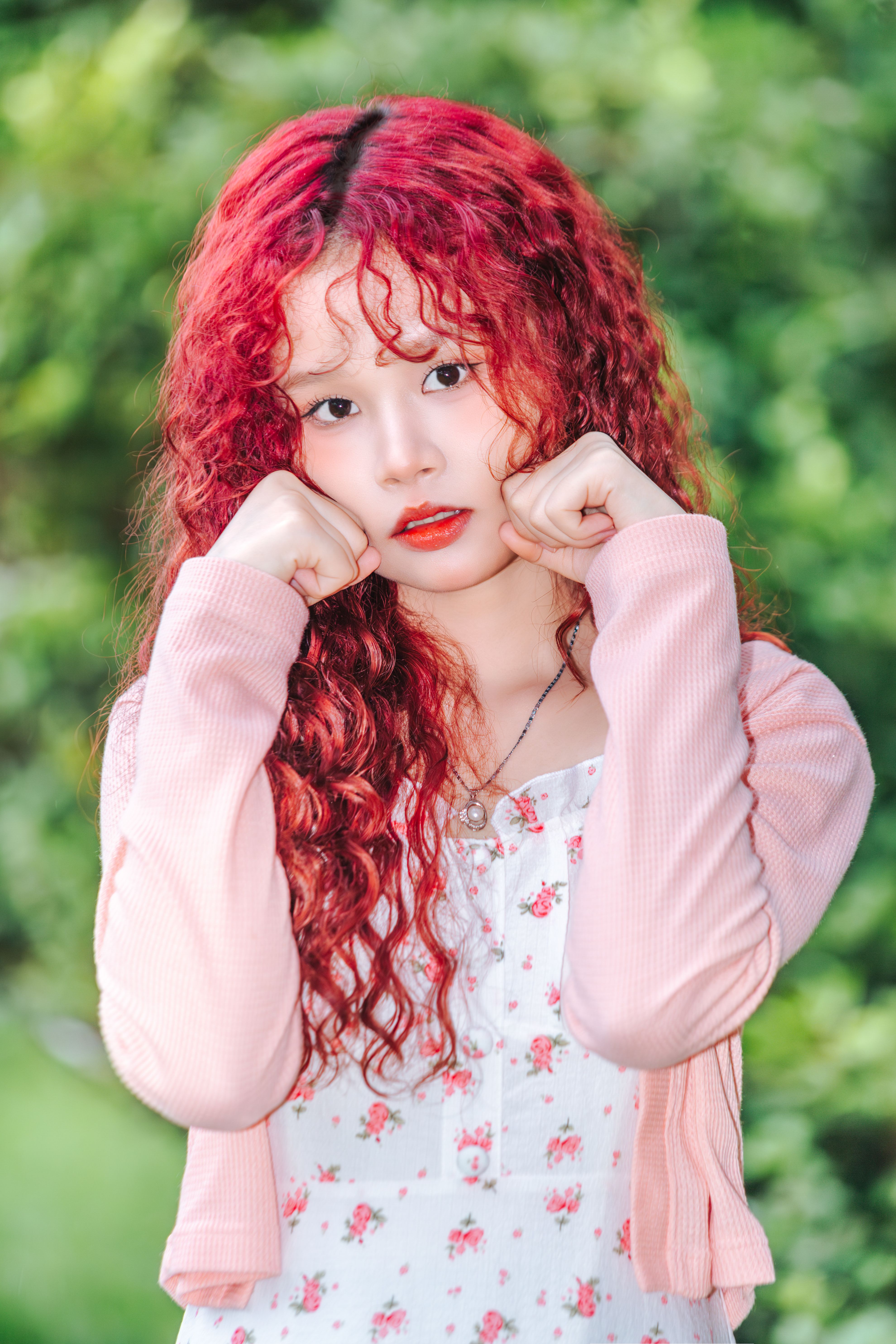 Portrait of a young woman with striking red hair posing outdoors against a lush green background.