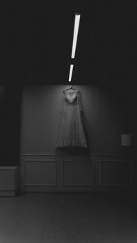 A black and white photo of a bridal gown hanging in a minimalist interior room.