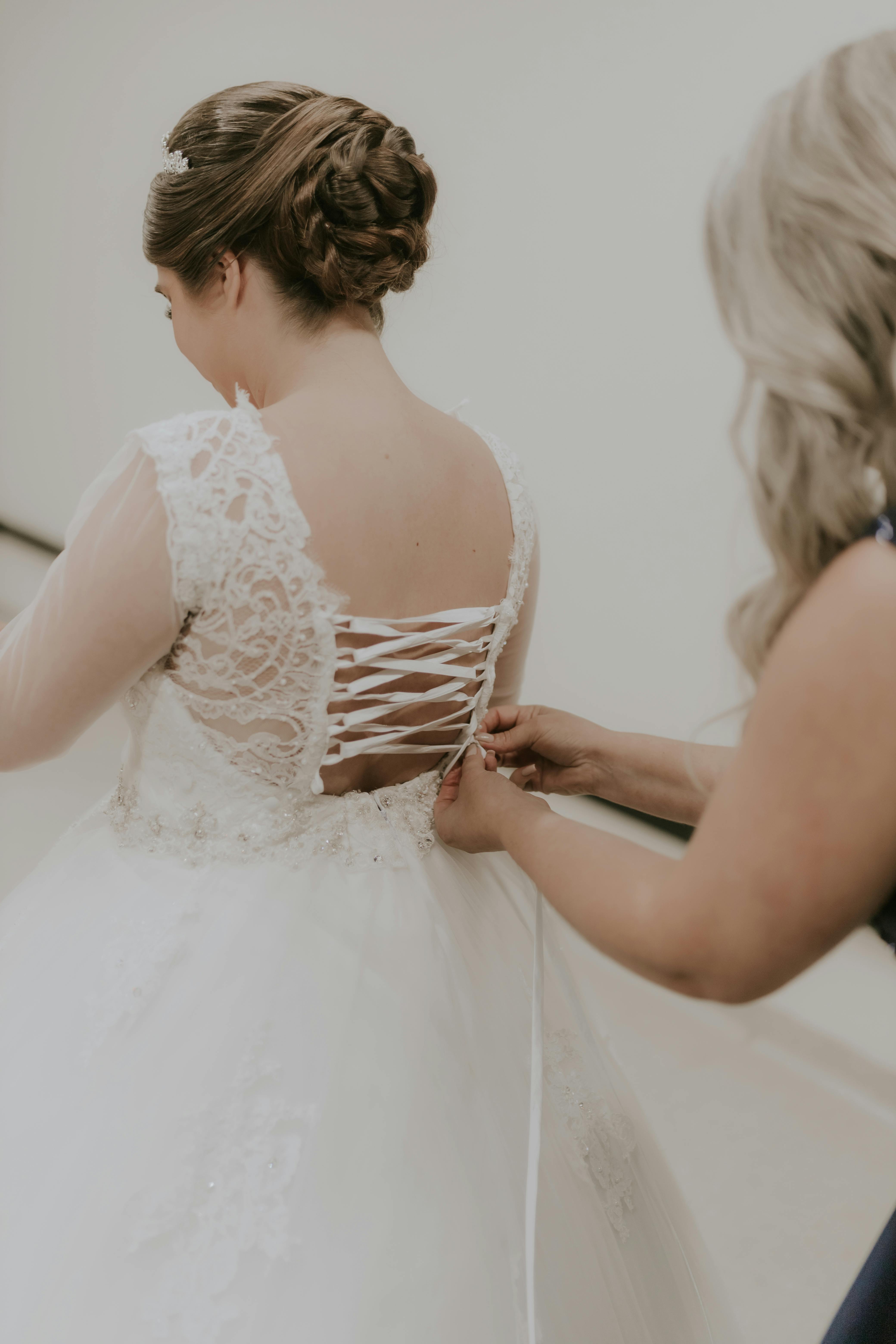 A bride in a lace gown getting ready with assistance indoors, capturing a cherished wedding moment.
