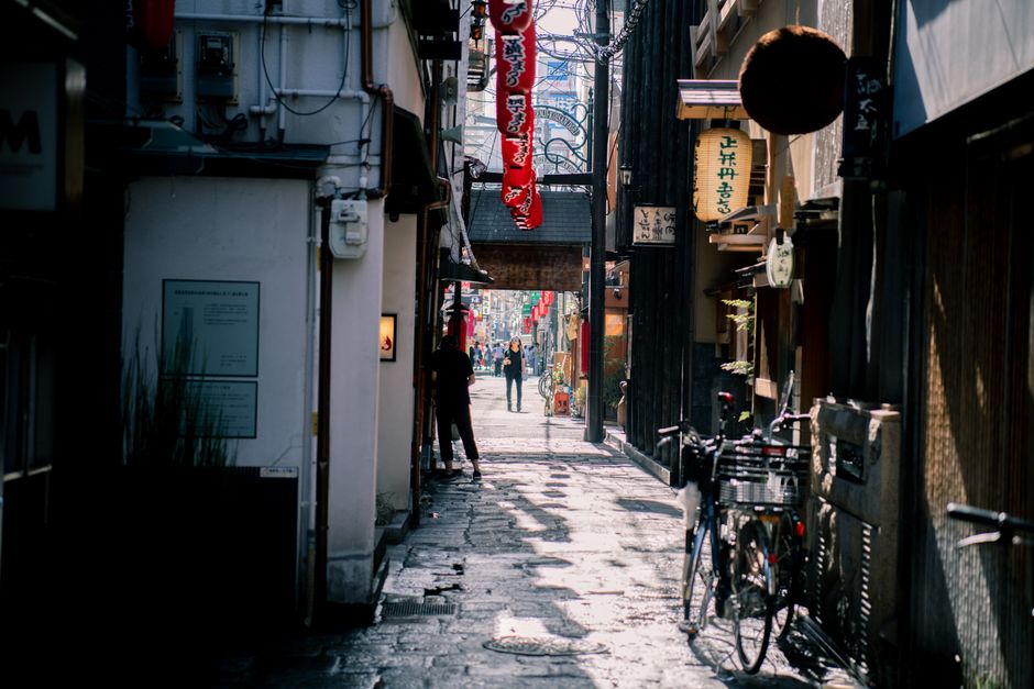 A narrow alleyway in Osaka, Japan featuring bicycles, people walking, and traditional signs. Captures urban street life.