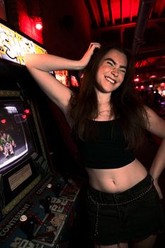 A joyful woman in a black top plays arcade games at night under vibrant lights.
