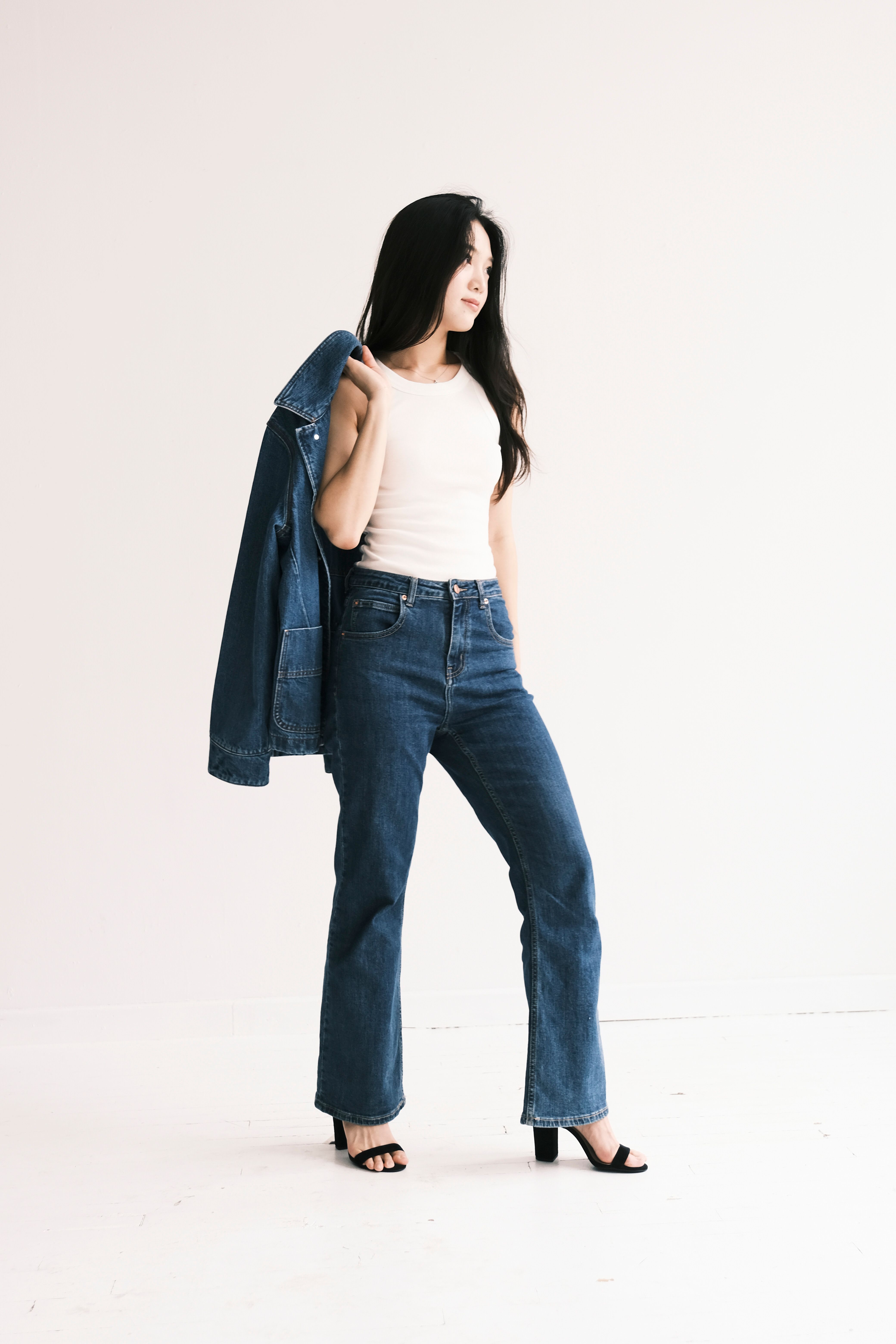 Young woman in fashionable denim holding jacket, posing confidently against a neutral backdrop.