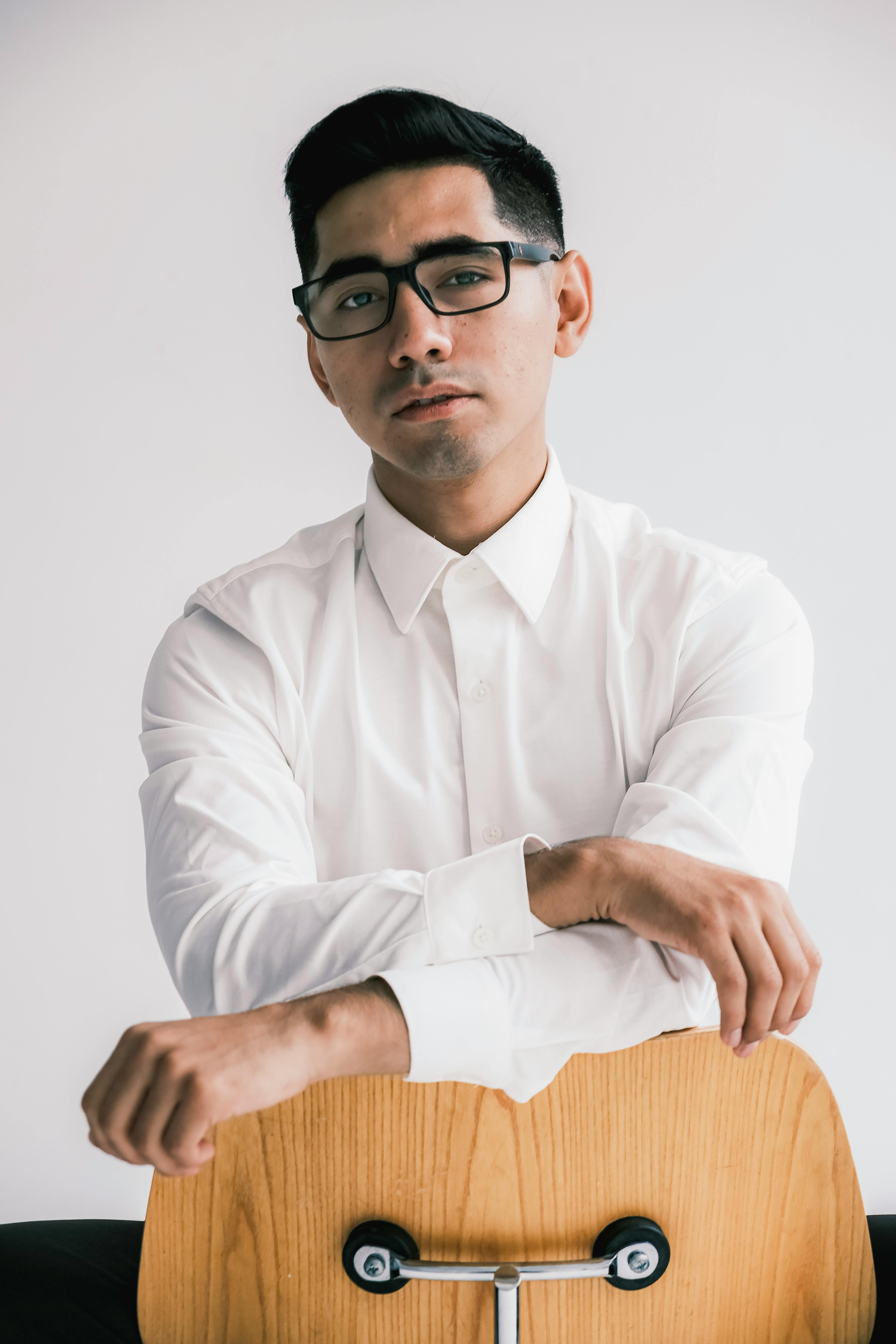 Portrait of a young man in a white shirt wearing glasses, sitting indoors with a confident expression.