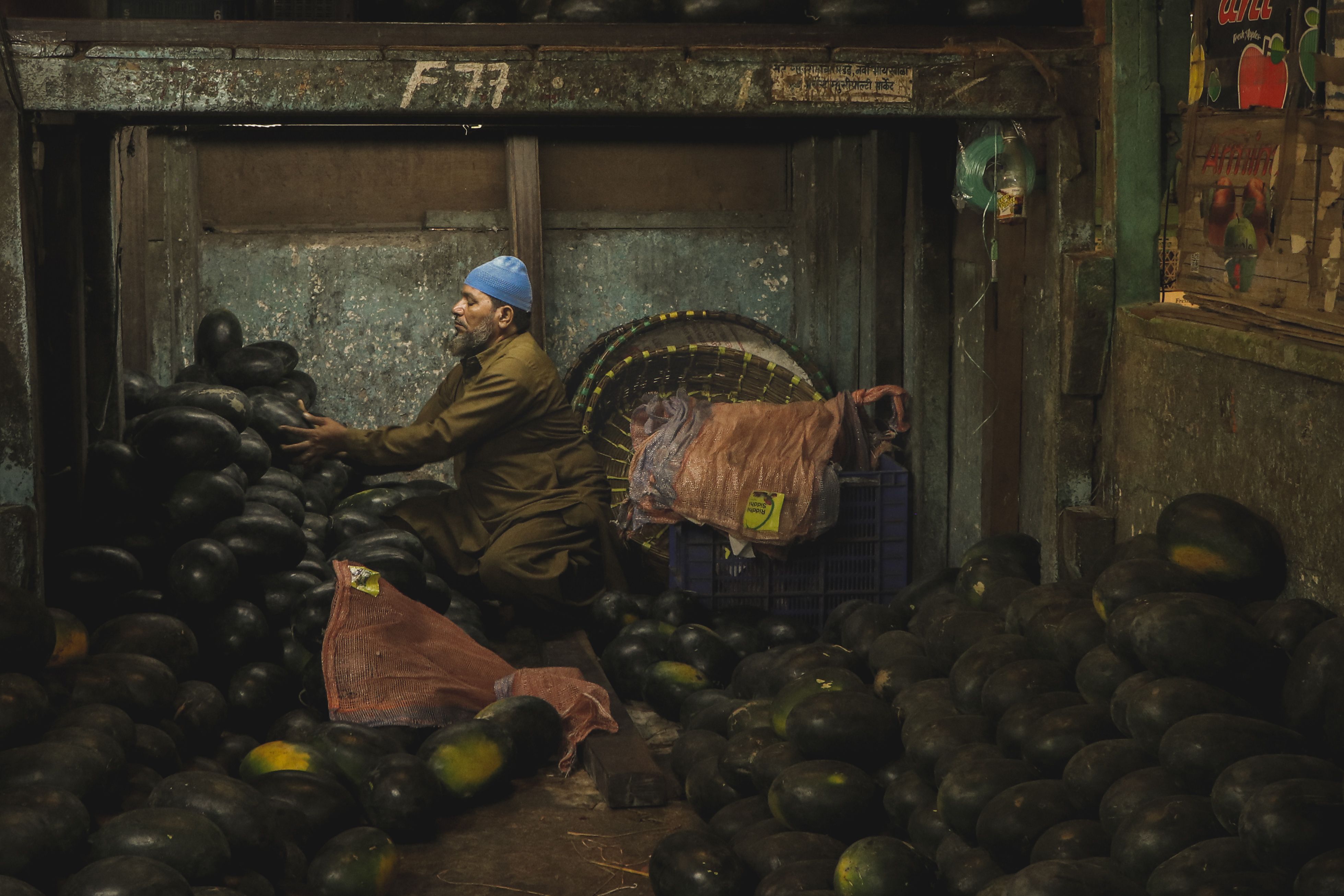 A man in a warehouse organizing a large quantity of black watermelons for market sale.