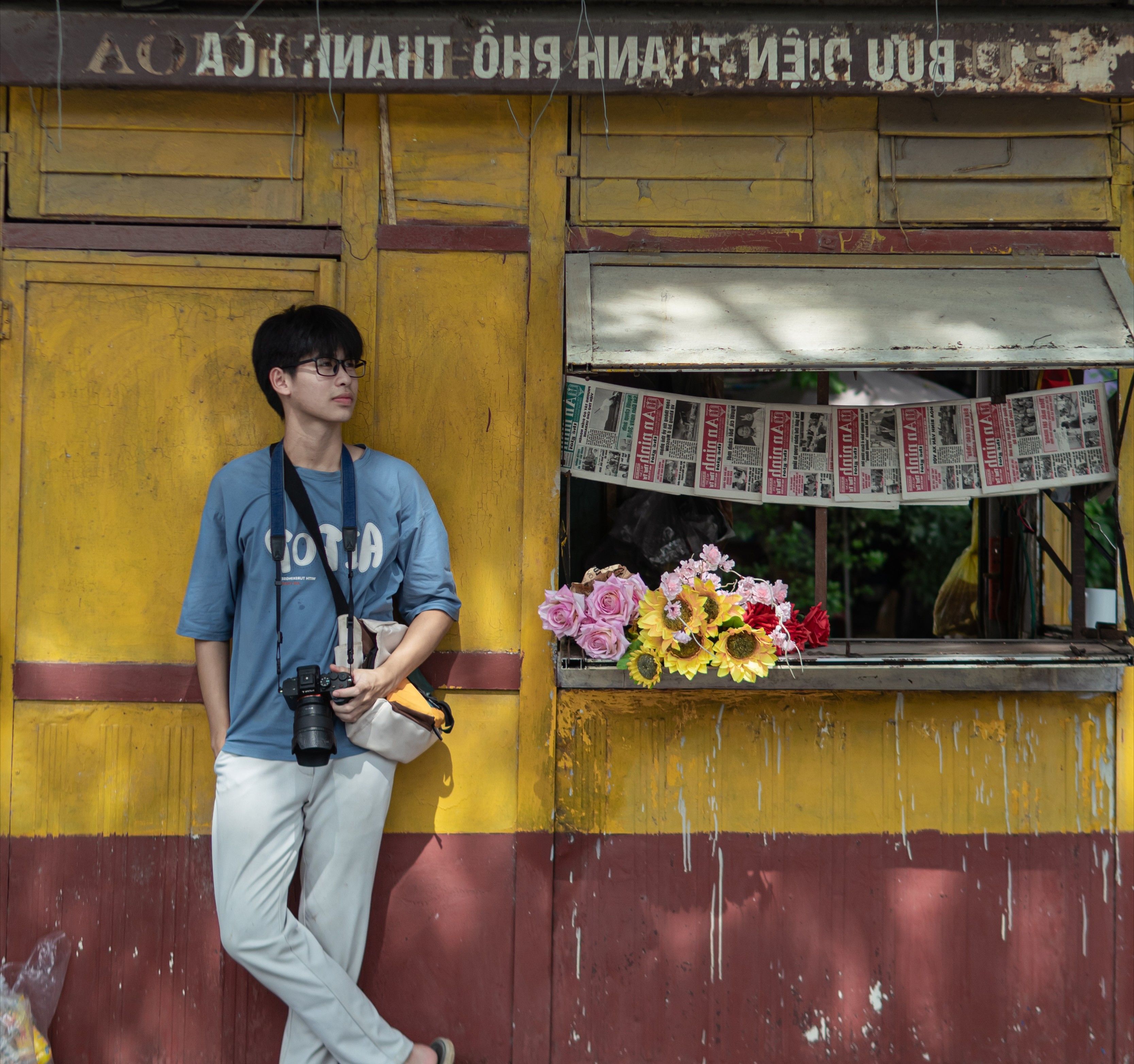 Young man with a camera leaning against a rustic yellow building with flowers.