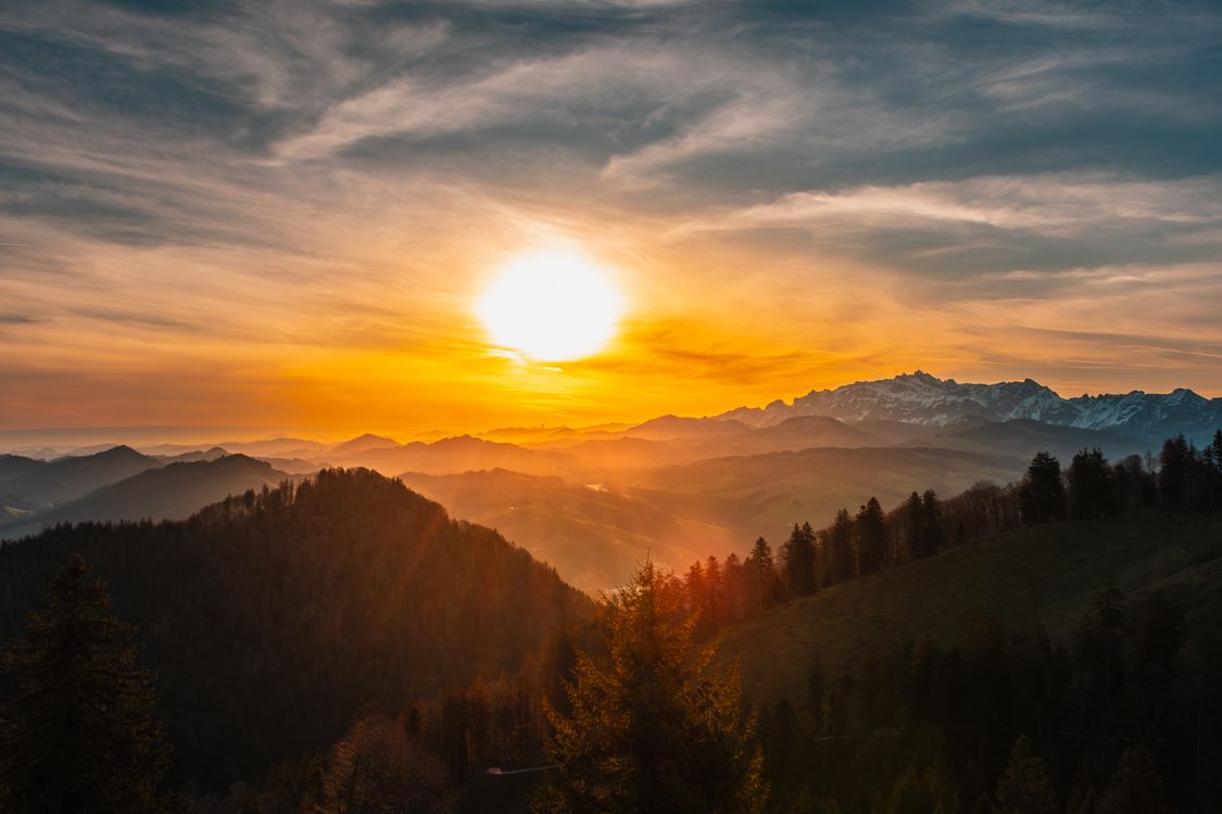 The sun sets over a mountain range with trees and mountains · Free