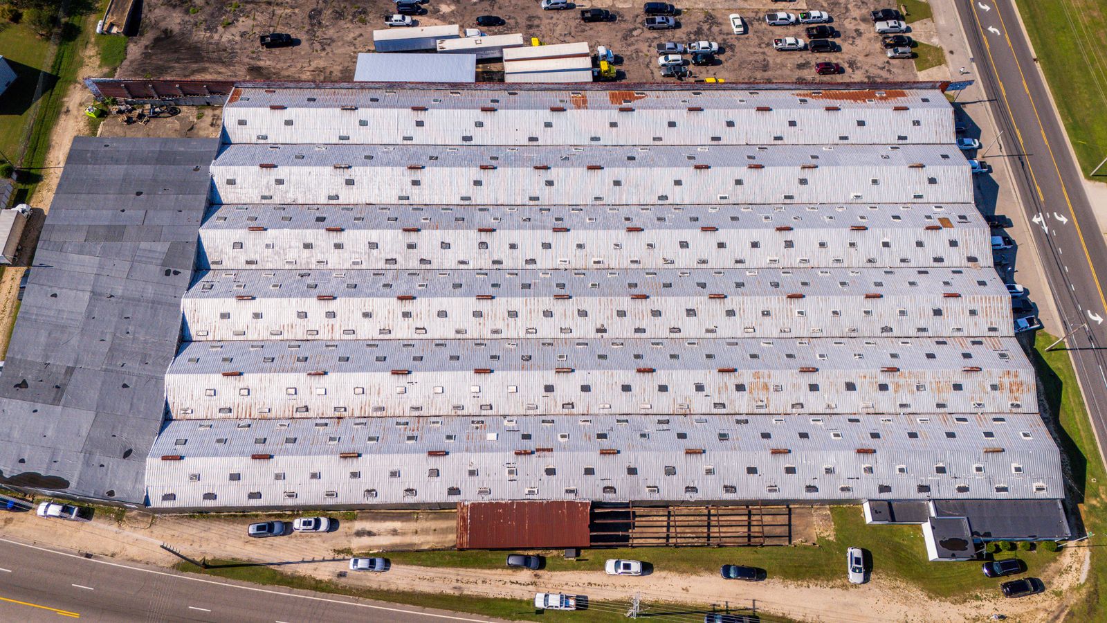 Aerial view of a long white-roofed warehouse beside a car park and trees