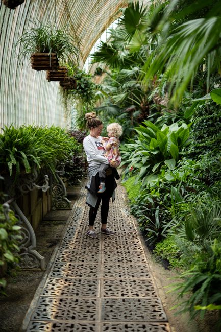 A woman carries her child through a lush, tropical greenhouse filled with vibrant flora.