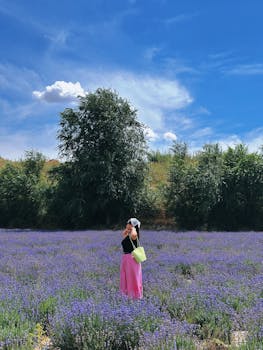 A woman in a lavender field under a blue sky, capturing nature's beauty in a rural Xinjiang, China setting.