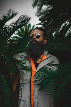 一位戴着墨镜、身穿夹克的时尚男士站在郁郁葱葱的棕榈叶中