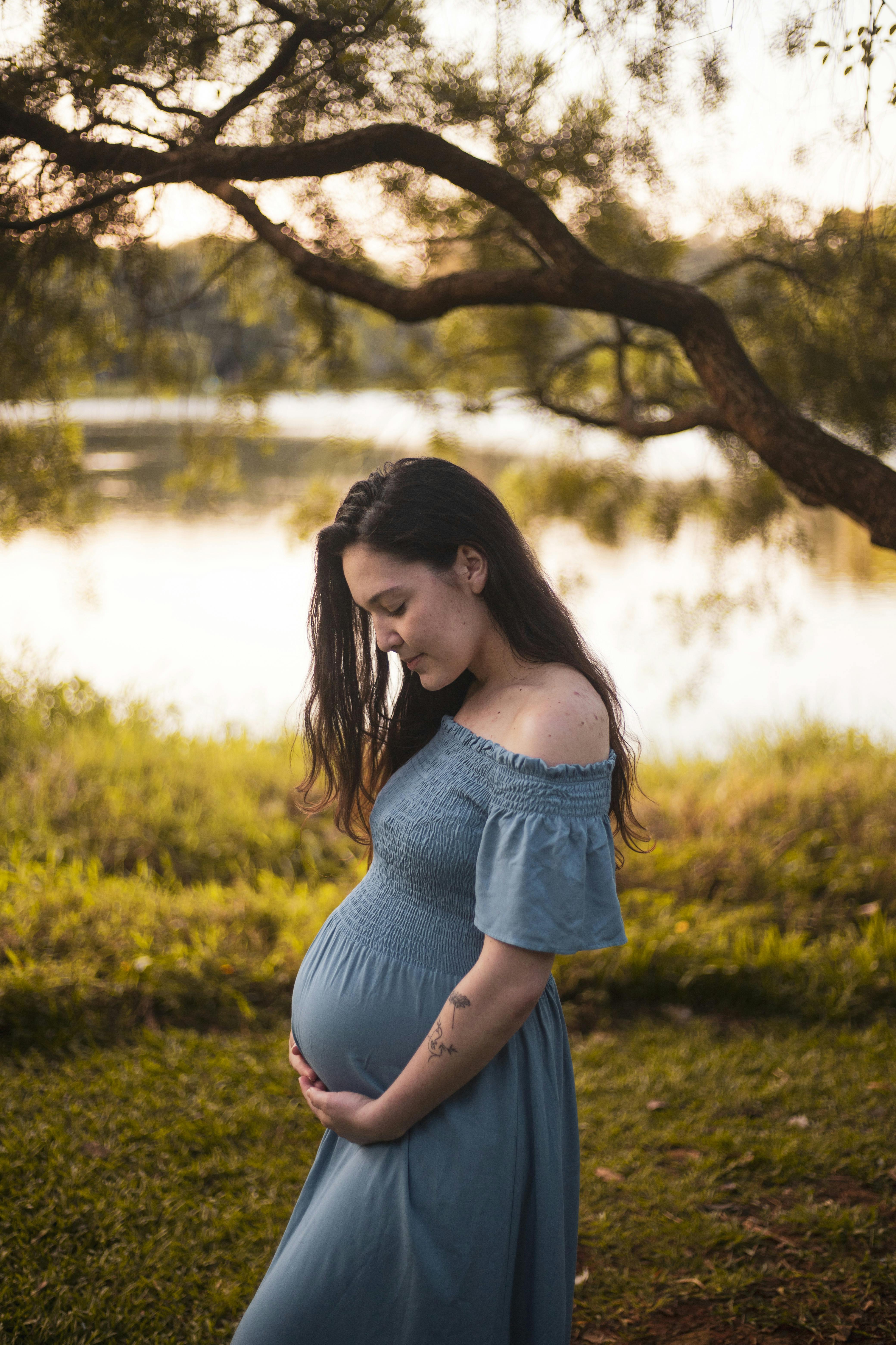 a pregnant woman in a blue dress stands by a lake