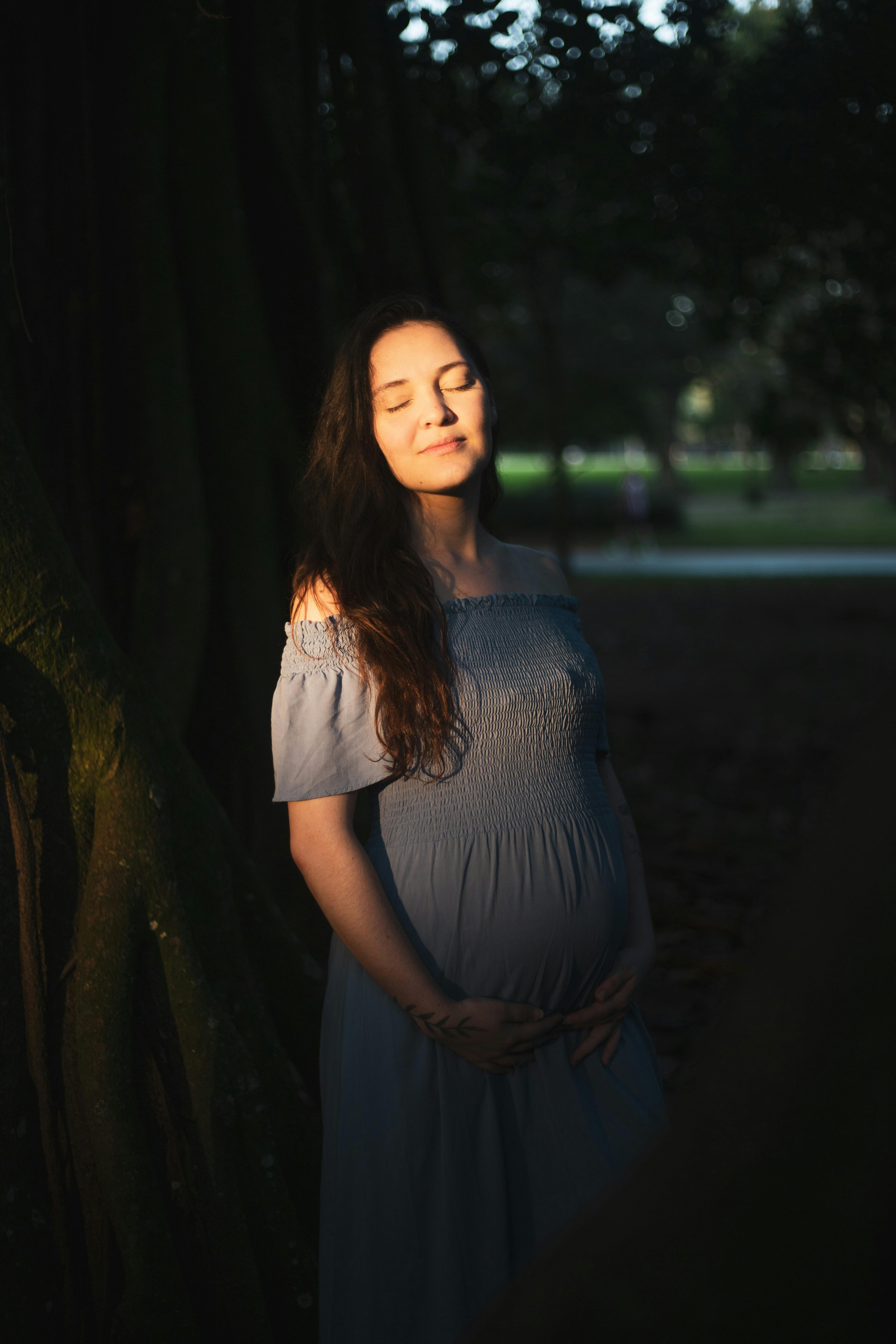 a pregnant woman in a blue dress stands in the shade