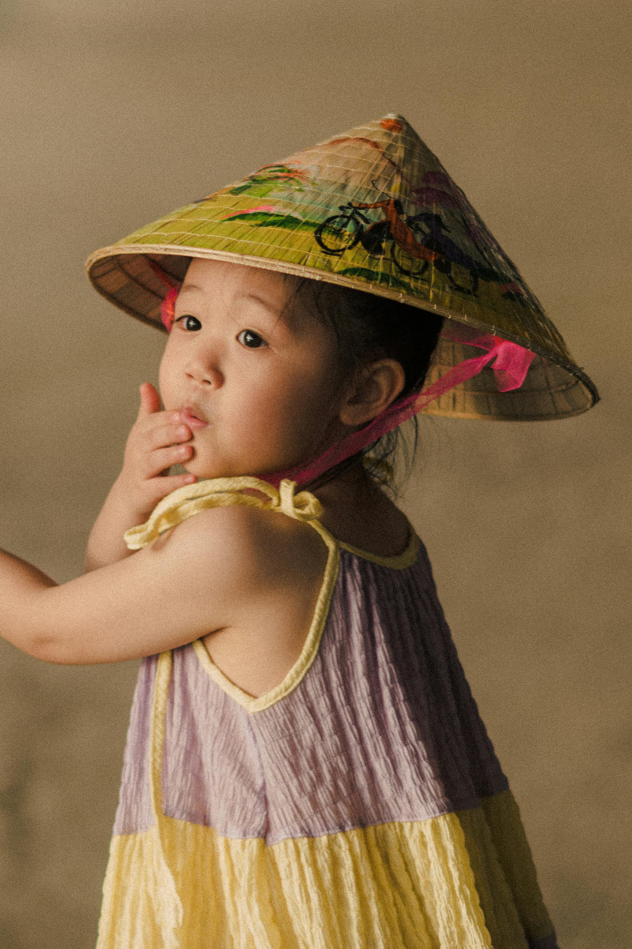 Cute Vietnamese child in a conical hat, expressing innocence and curiosity, outdoors in summer.