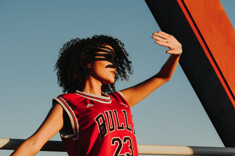 A young woman in a sports jersey shields her eyes from the setting sun, casting dramatic shadows.