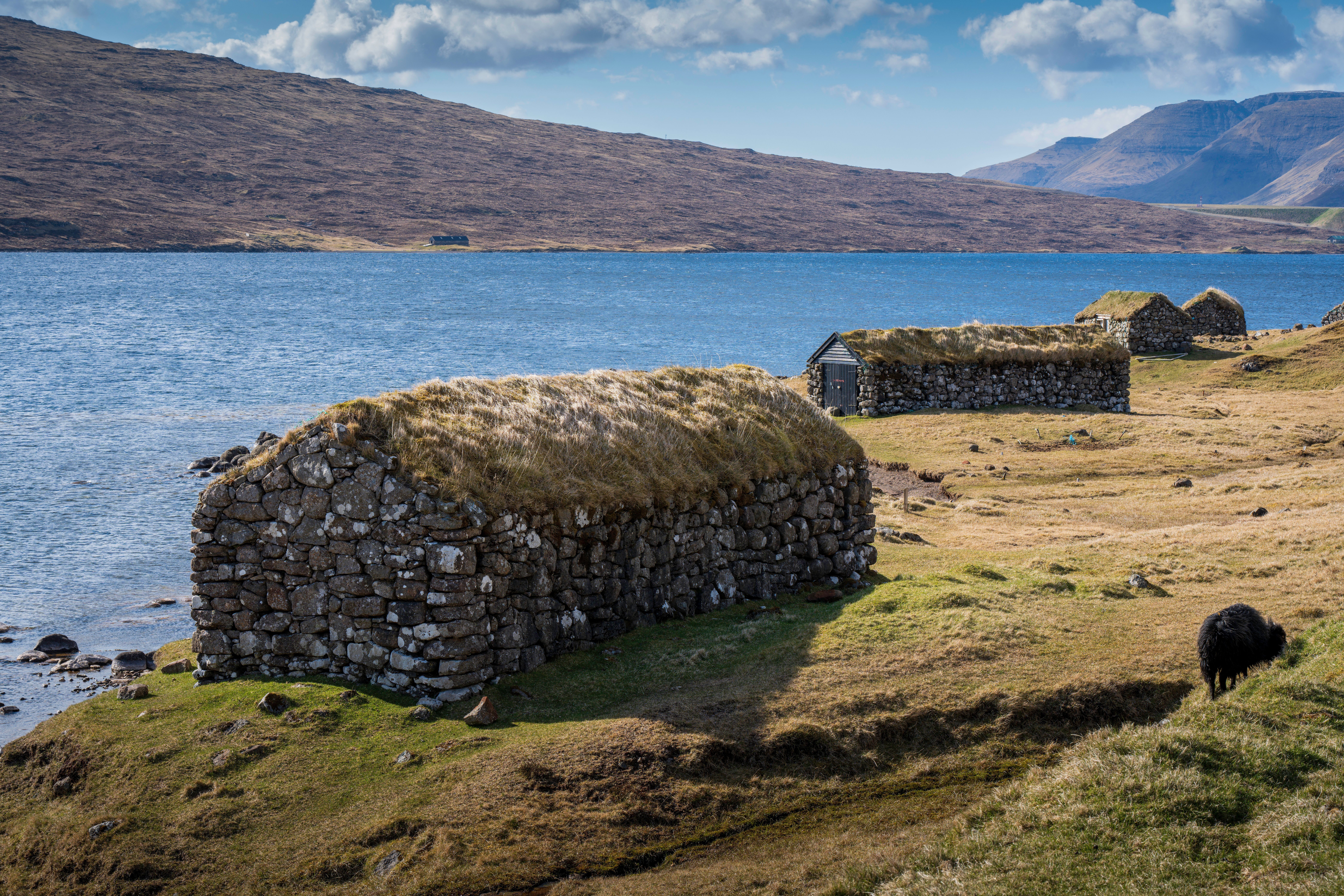 Scenic view of turf-roofed stone houses by a tranquil lake in the Faroe Islands, surrounded by mountains.