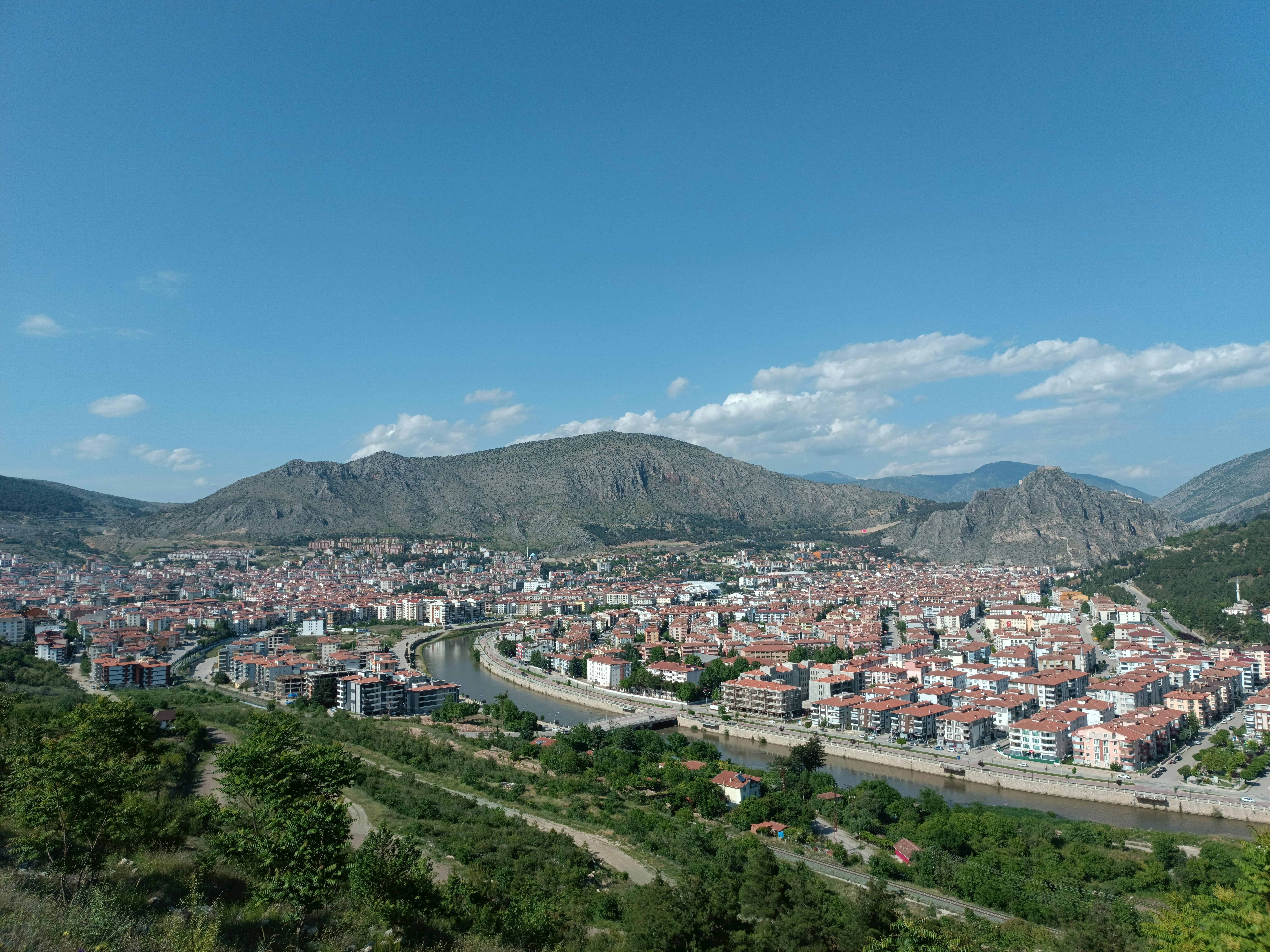 A panoramic view of a city nestled between lush greenery and rugged mountains under a clear blue sky.