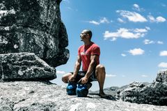 A muscular man exercises with kettlebells on rocky ground under a bright blue sky, emphasizing strength and fitness.