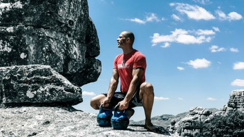 A muscular man exercises with kettlebells on rocky ground under a bright blue sky, emphasizing strength and fitness.