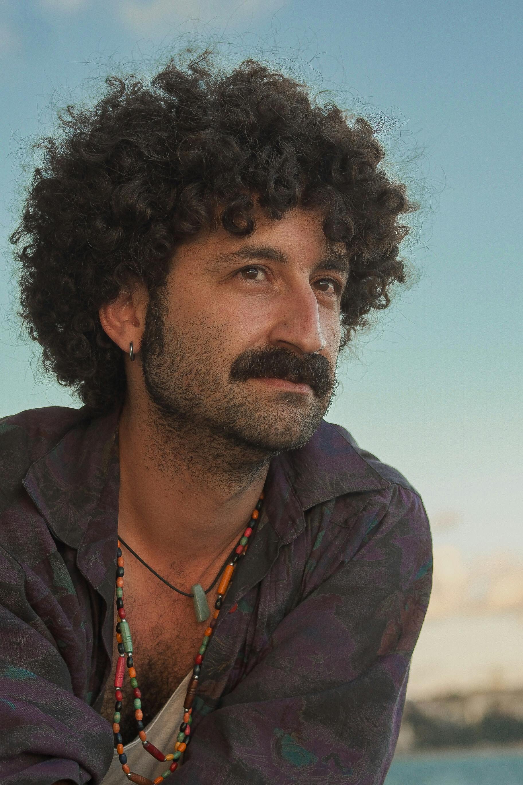 A thoughtful adult man with a mustache and curly hair outdoors, wearing a colorful necklace and shirt.