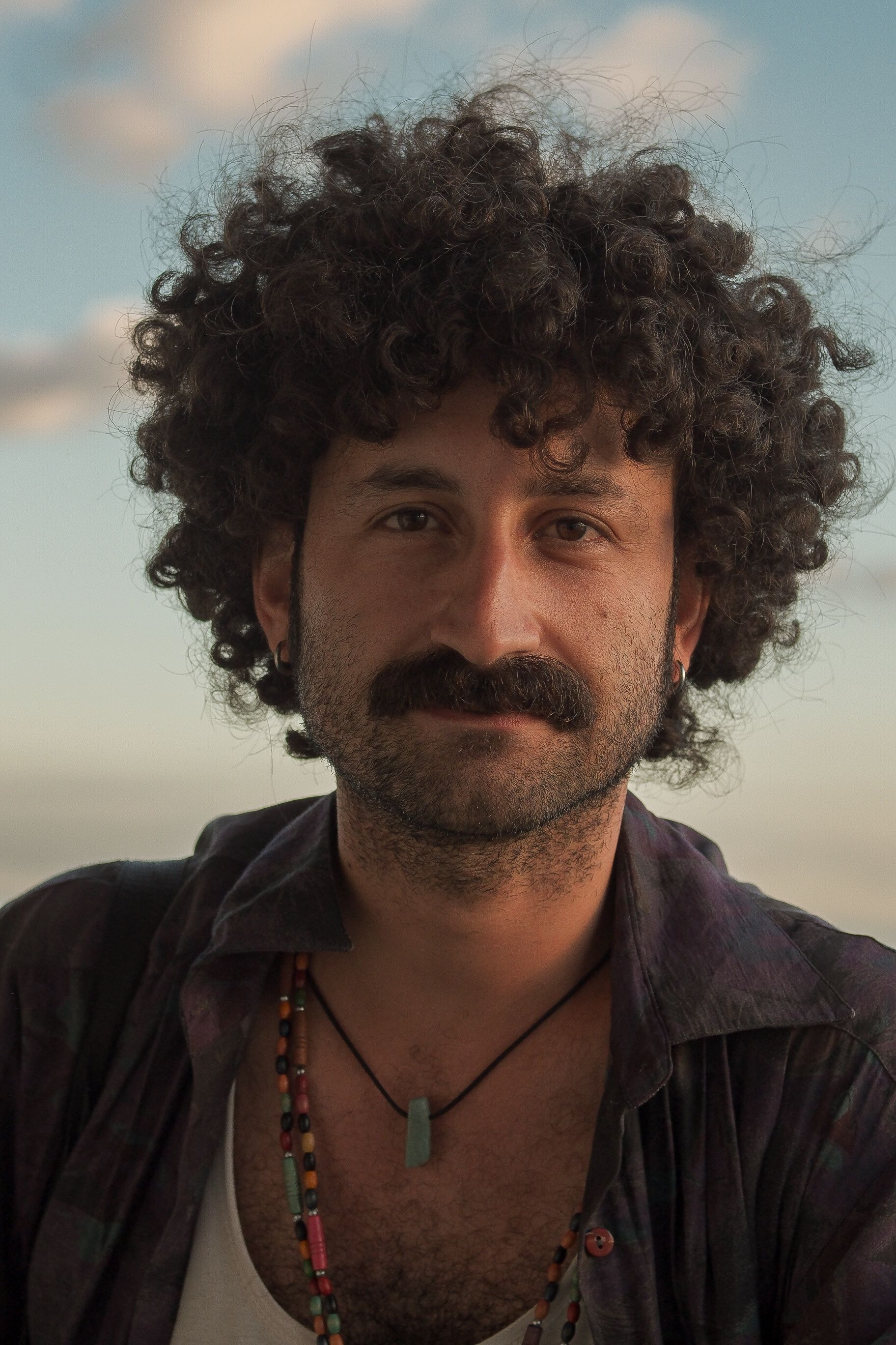 Casual outdoor portrait of a man with curly hair and beaded necklace at dusk.