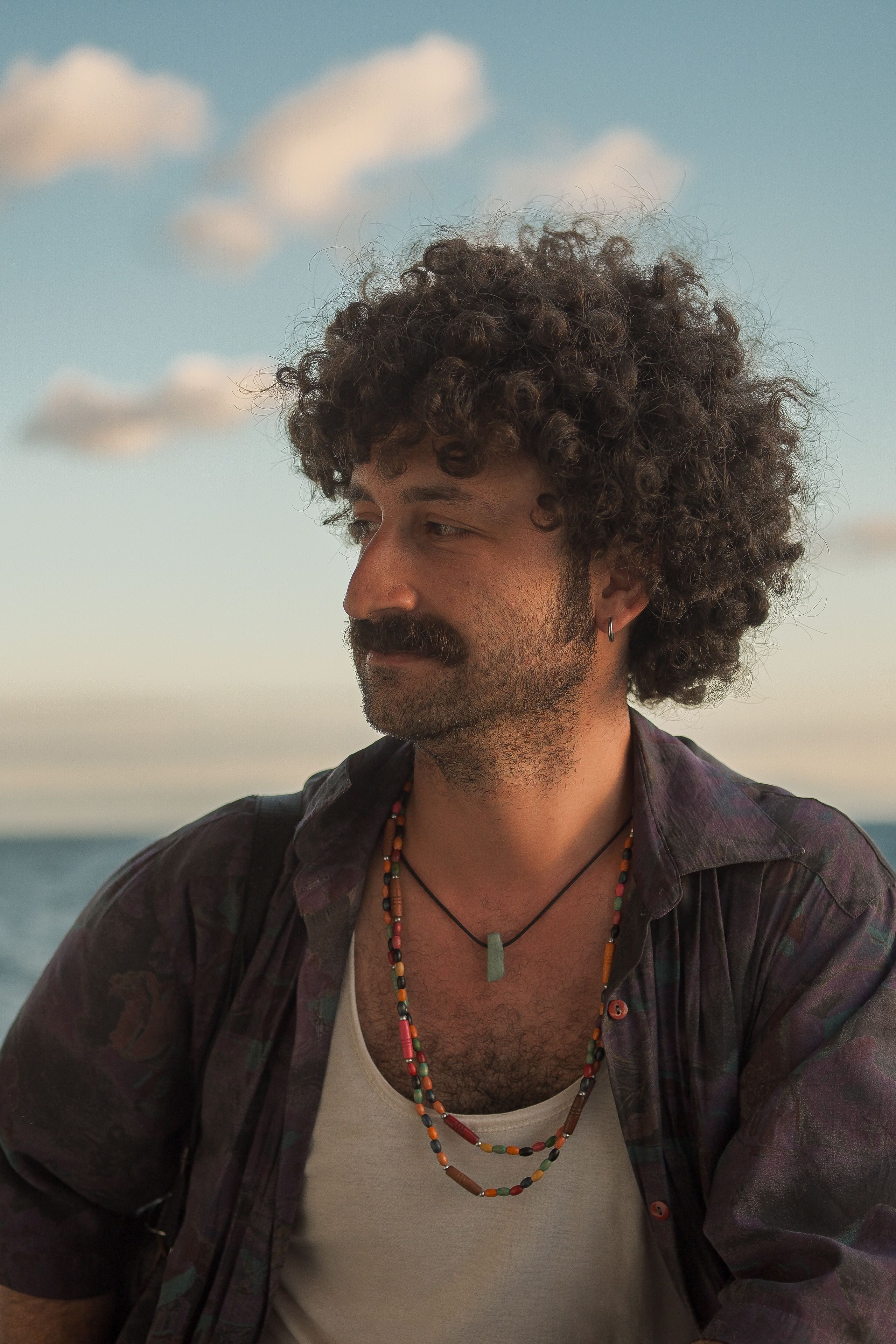 A stylish man with curly hair and a necklace, gazing at the beach during sunset.