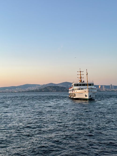 Free A ferry cruises on a calm ocean with a city skyline at sunset, offering a serene travel experience. Stock Photo