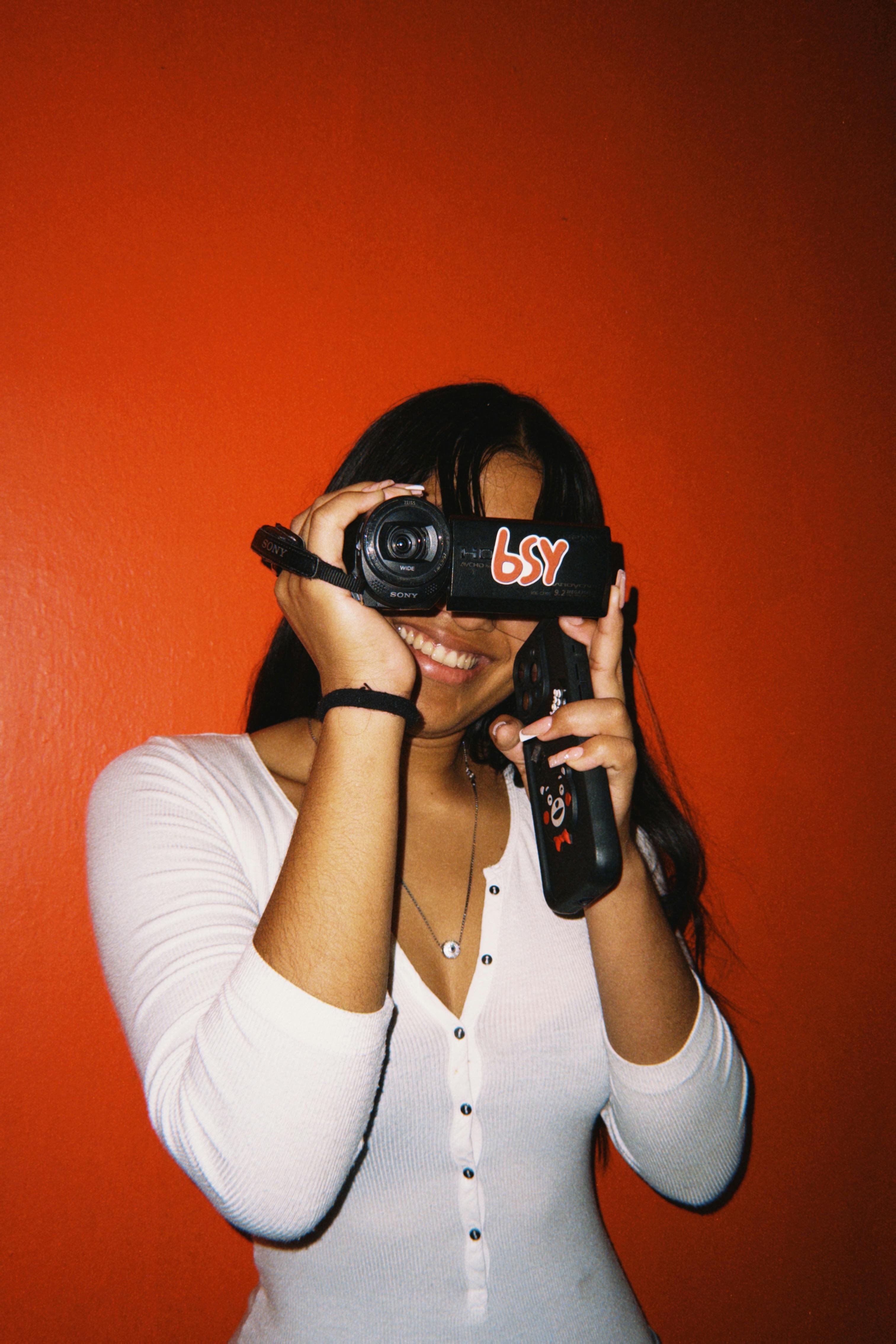 Smiling young woman capturing a moment with a camcorder against a bold orange background.