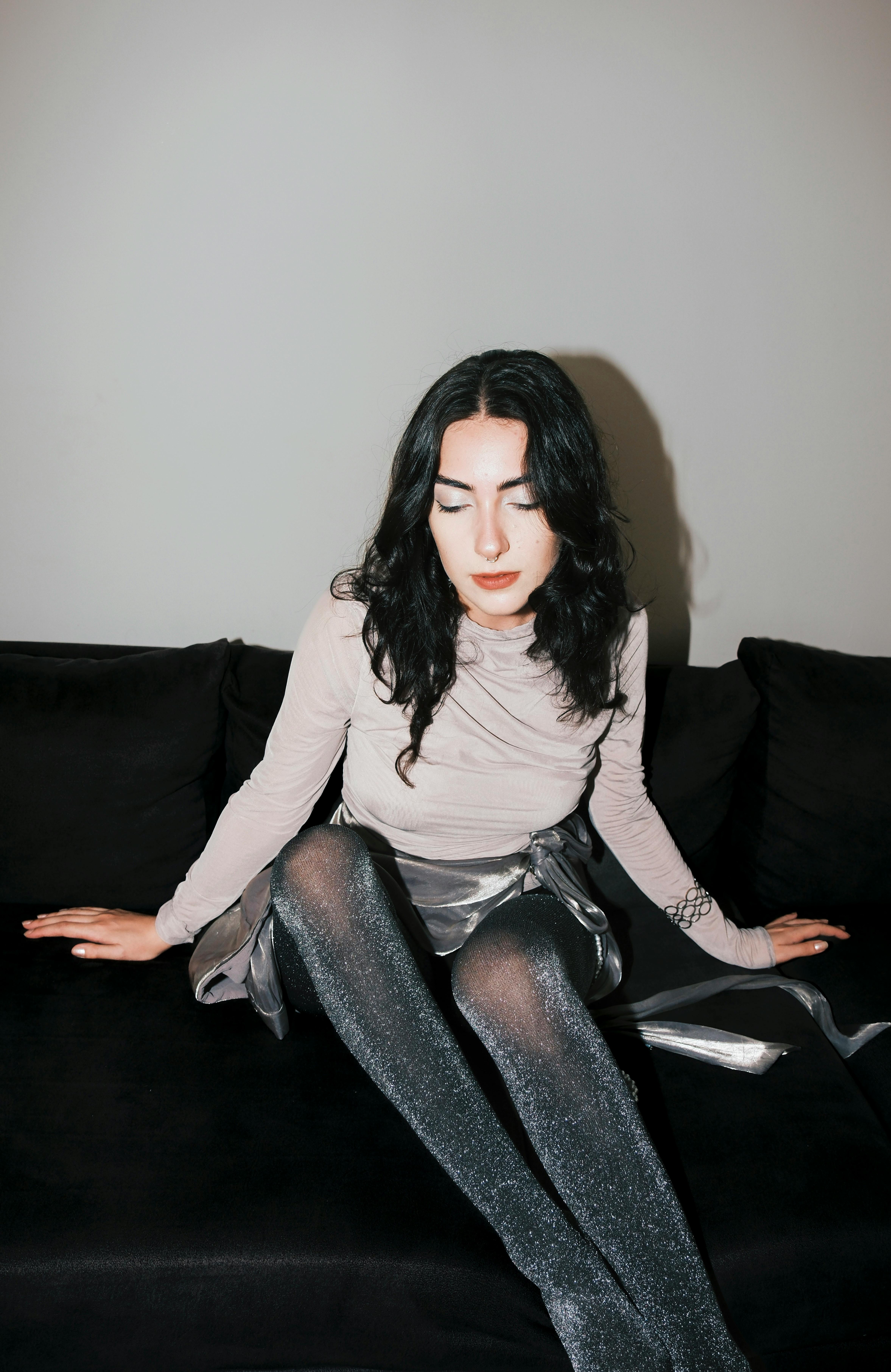 A young woman in fashionable metallic attire sits gracefully indoors on a dark couch.