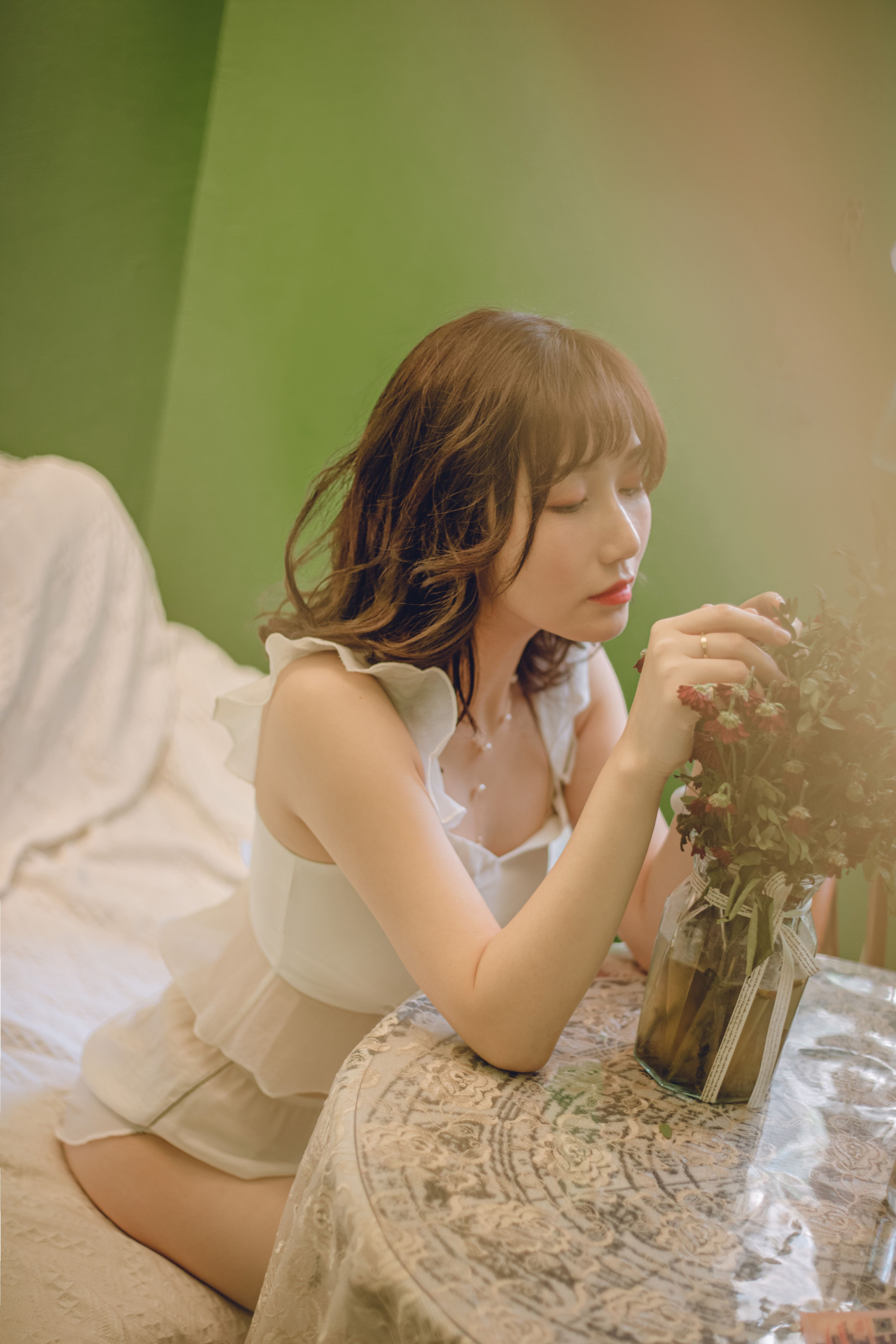 A woman in a white dress sits peacefully at a table with flowers, capturing an elegant and serene moment indoors.