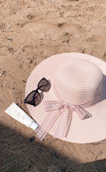 Stylish pink sun hat with sunglasses and sunscreen on a sandy beach, perfect for summer relaxation.