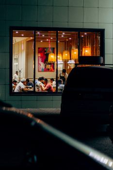 A warm view of a bustling restaurant interior seen through a window at night, with people dining inside.