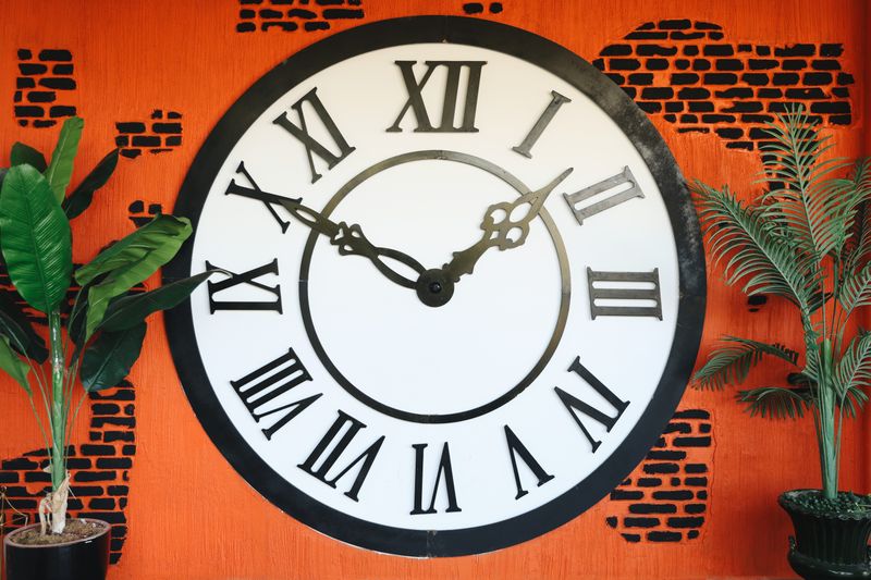 A vintage-style wall clock with Roman numerals against a vibrant orange brick background.