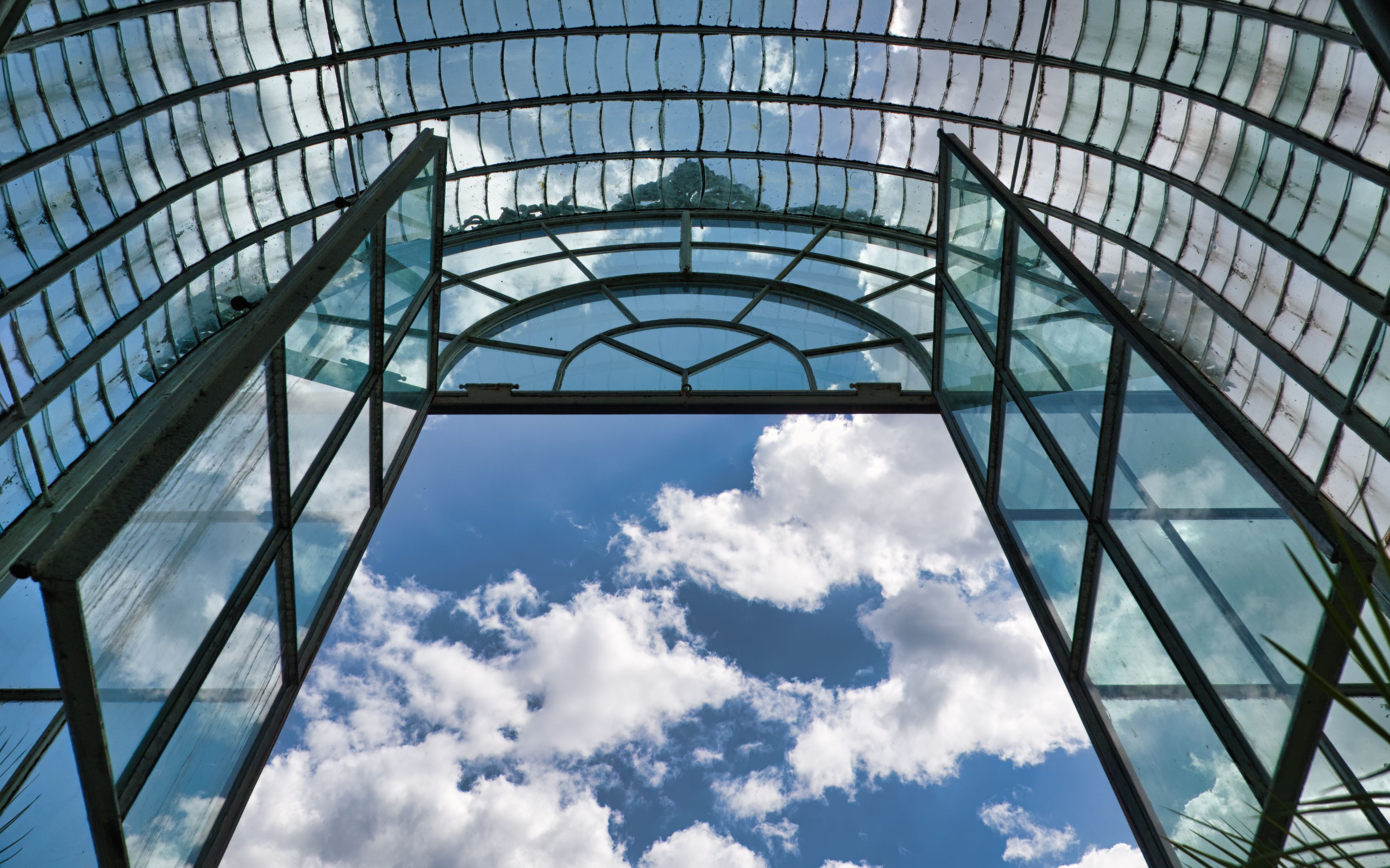 Glass arched roof of a greenhouse against a blue sky with clouds, showcasing modern architecture.